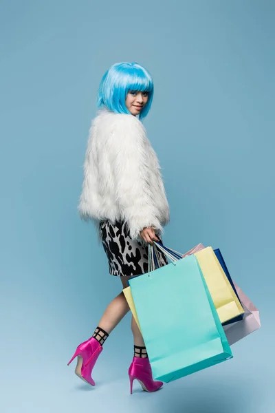 Elegante mulher asiática em peruca e casaco fofo segurando sacos de compras e sorrindo para a câmera no fundo azul — Fotografia de Stock
