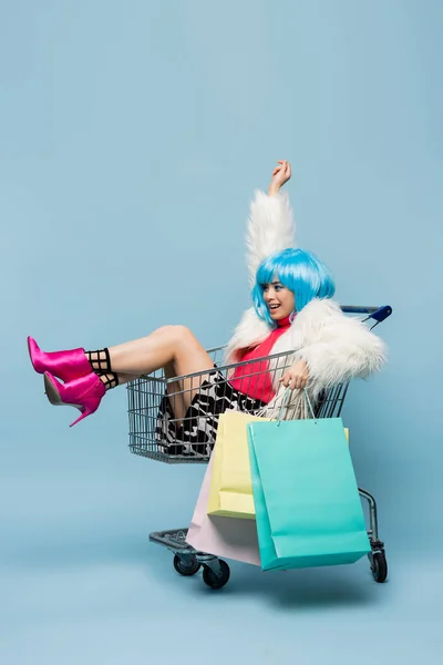Alegre asiático pop arte mujer holding compras bolsas mientras sentado en carro en azul fondo - foto de stock