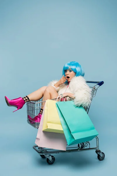 Erstaunte asiatische Frau im Pop-Art-Stil schaut in die Kamera, während sie Einkaufstüten im Einkaufswagen auf blauem Hintergrund hält — Stockfoto