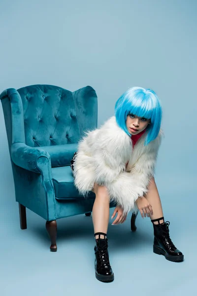 Asian woman in bright wig and fluffy jacket looking at camera on turquoise armchair on blue background — Stock Photo
