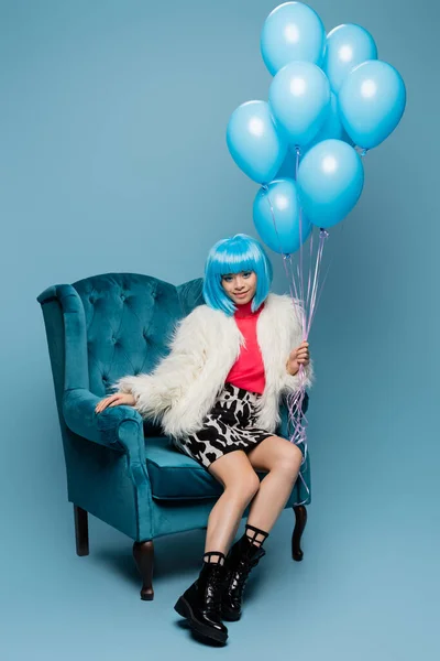 Улыбающаяся азиатская поп-арт женщина держит воздушные шары на кресле на синем фоне — стоковое фото