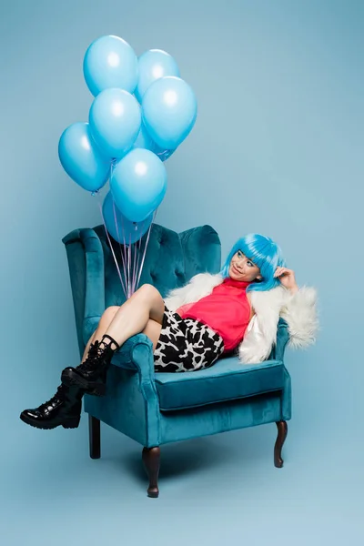 Happy asian model in pop art style looking at balloons on armchair on blue background — Stock Photo