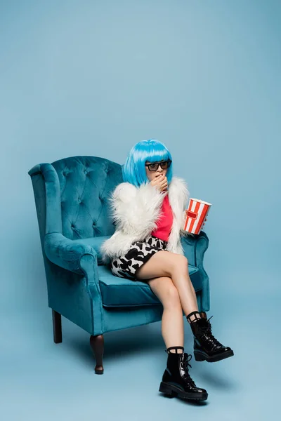 Elegante asiático pop arte mujer en gafas de sol comer palomitas de maíz en sillón sobre fondo azul - foto de stock