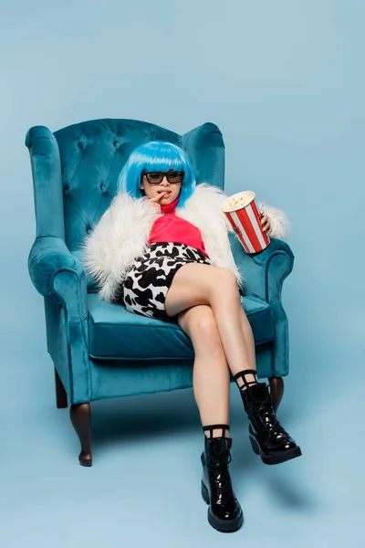 Stylish asian woman in fluffy jacket and sunglasses holding popcorn in armchair on blue background — Stock Photo