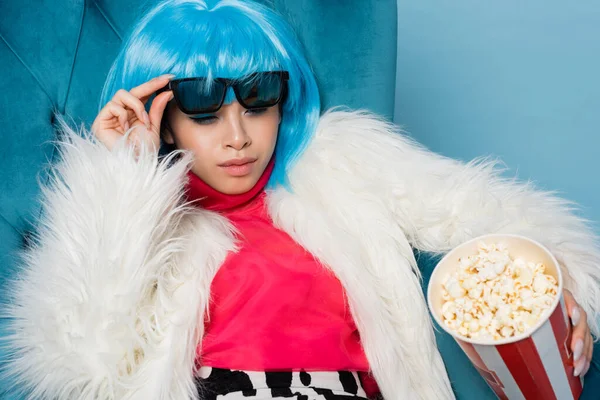 Pretty asian woman in fluffy jacket holding popcorn and sunglasses in armchair isolated on blue — Stock Photo