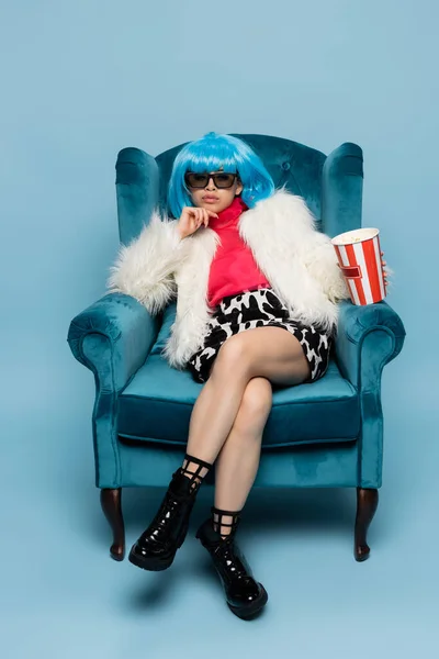 Азиатка в поп-арте держит попкорн, сидя на бархатном кресле на синем фоне — стоковое фото