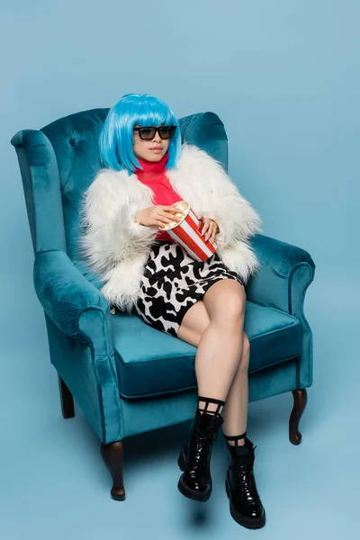 Modelo asiático en estilo pop art sosteniendo palomitas de maíz en sillón sobre fondo azul - foto de stock