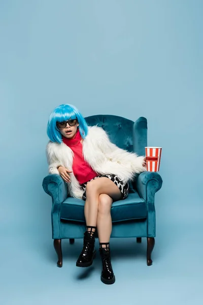 Amazed asian pop art woman in sunglasses holding popcorn on armchair on blue background — Stock Photo