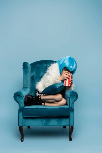Испуганная азиатская поп-арт женщина в ярком парике держит попкорн на кресле на синем фоне — стоковое фото