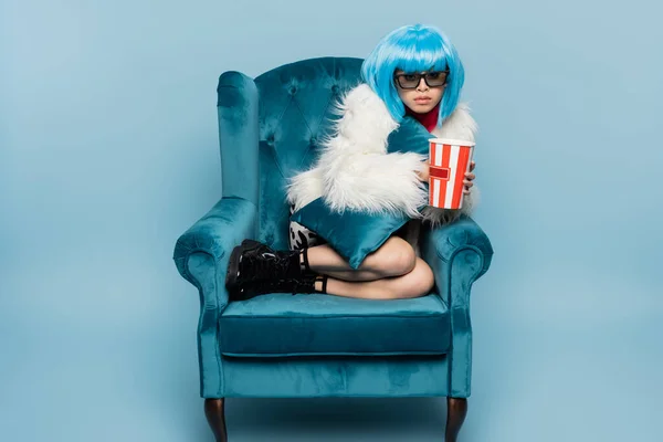 Asian woman in pop art style holding popcorn and pillow on armchair on blue background — Stock Photo