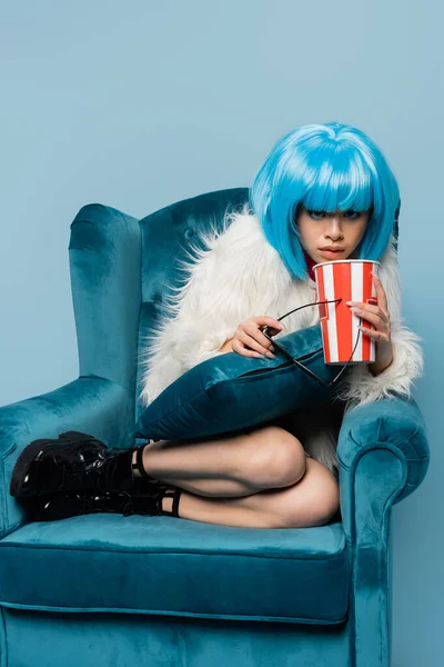 Stylish asian pop art woman holding sunglasses and popcorn on armchair isolated on blue — Stock Photo