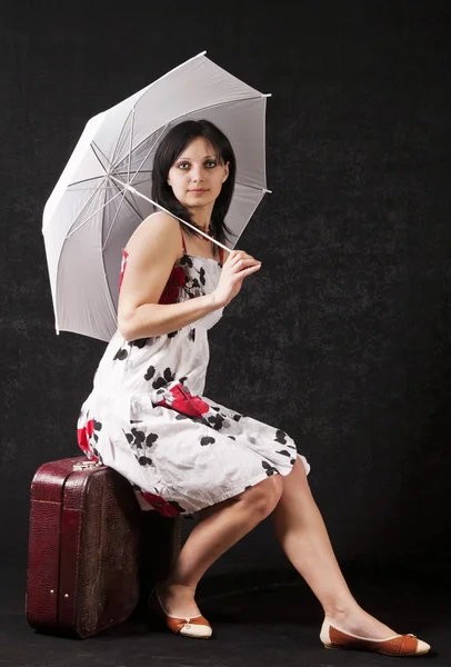 Donna con un ombrello in abito estivo in posa in studio seduta su una valigia — Foto Stock