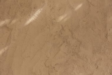 Yumuşak doğal ışık ve ince gölgelerle aydınlatılmış bej alçı duvar dokusunun yakın çekimi. Tasarım, mimari ve doku referansları için minimal ve sıcak yüzey arkaplanı ideali.