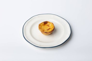 Üç geleneksel Portekiz kremalı turtasına (pastis de nata) yakın plan, temiz beyaz arka plana yerleştirilmiş, mavi kenarlı beyaz bir tabağa yerleştirilmiş. Süslü altın kabuk ve karamelize krema. Baştan çıkarıcı ve otantik bir görünüm yaratır.