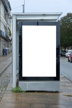 Porto, Portekiz 'de yağmurlu bir caddedeki otobüs durağında boş reklam panosu ıslak kaldırımlar, park edilmiş arabalar ve arka planda şemsiyelerle yürüyen insanlar..