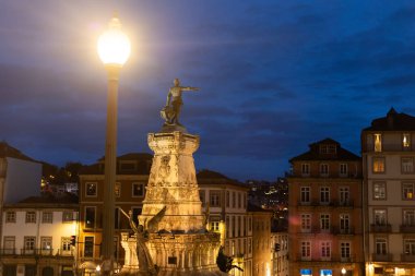 Porto, Portekiz 'deki tarihi bir anıtın akşam manzarası. Derin mavi gökyüzünün altındaki sıcak sokak lambalarıyla aydınlatılmış. Heykel, geleneksel binalarla çevrili taş bir kaidede duruyor.