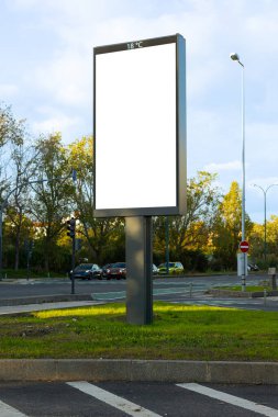 Arka planda ağaçlar ve arabalar olan bir cadde kavşağında dikey boş ilan panosu duruyor. Boş beyaz reklam panosu, açık bir gökyüzü ile doğal gün ışığında çekilen modelleme ya da ticari tasarım yerleştirme için hazır.