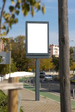 Güneşli bir öğleden sonra ağaçlar ve konut binalarıyla çevrili sessiz bir şehir kaldırımında dikey boş bir ilan panosu..