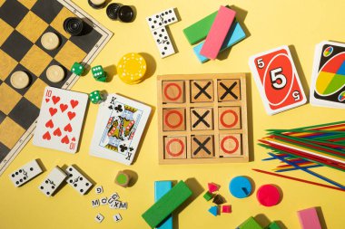 Sarı arka planda, tic-tac-toe tahtası, iskambil kartları, domino, zar, UNO kartları, kaldırma çubukları, poker fişleri, dama taşları ve renkli tahta bloklar da dahil olmak üzere çeşitli masa üstü oyunlarının düz kompozisyonu. Neşeli ve enerjik.
