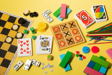 Sarı arka planda, tic-tac-toe tahtası, iskambil kartları, domino, zar, UNO kartları, kaldırma çubukları, poker fişleri, dama taşları ve renkli tahta bloklar da dahil olmak üzere çeşitli masa üstü oyunlarının düz kompozisyonu. Neşeli ve enerjik.