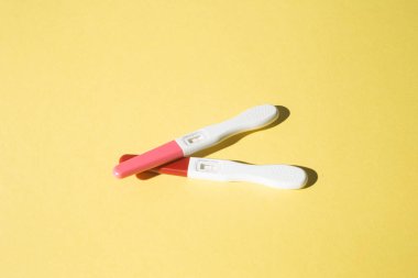 İki hamilelik testi yumuşak sarı arka planda yatar, temiz ve minimum bir kompozisyonda düzenlenmiştir. Doğrudan ışık, düz yüzeyleri ve nesnelerin basit tasarımını vurgulayarak tanımlanmış gölgeler yaratır. Sahne bir beklenti hissi veriyor.