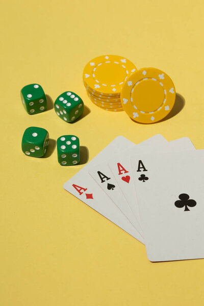 Flat lay of casino elements on a yellow background, featuring green dice, yellow poker chips, and a winning hand of four aces. The composition conveys themes of gambling, luck, and gaming in a clean and minimal style.