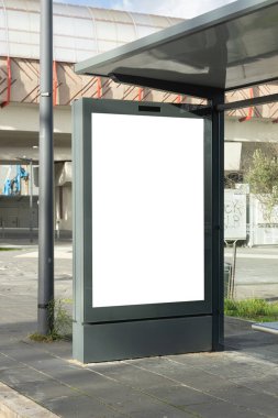 Modern bir otobüs durağındaki dikey reklam panosunda boş beyaz bir ekranın özel içerik için hazır olduğu Mockup Blank reklam panosu. Sahne, kaldırımlı bir şehir ortamında.