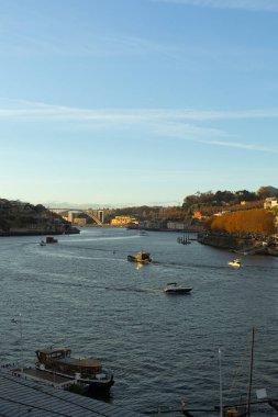 Portekiz 'in Porto kentindeki Douro Nehri' nin sakin sularda yüzen teknelerle çekilmiş manzarası. Şehir silueti, tarihi nehir kenarı binaları ve çevreleyen tepeler berrak mavi gökyüzünün altında huzurlu bir şehir manzarası yaratıyor.