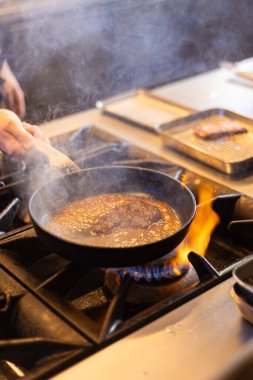 Profesyonel bir mutfakta, açık bir gaz alevinin üzerinde biftek pişiriyorum. Et, görünür bir buhar ve alevlerle zengin bir sosta kızarıyor ve dinamik, dramatik bir pişirme atmosferi yaratıyor. Bu resim profesyonel gastronomiyi temsil ediyor.