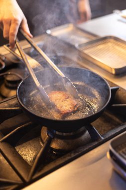 Profesyonel bir mutfağın içinde bir et parçasını kızgın tavada yakan bir şefin yakın çekimi. Et pişerken tavadan buhar yükseliyor. Restoran ortamında yemek hazırlama ve ısıyı vurguluyor..