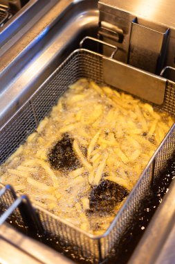 Profesyonel mutfak fritözü ve kızgın yağda kızaran patates kızartmasıyla dolu metal bir sepet. Ticari paslanmaz çelik fritözün yakın plan görüntüsü, kızartma işlemini gösteriyor