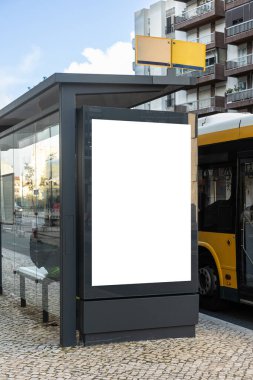 Kentsel bir caddedeki modern bir otobüs durağındaki boş dikey reklam panosu. Boş beyaz ekran, kaldırım taşları ve konut binalarıyla çevrili sarı bir belediye otobüsünün yanında duruyor..