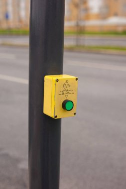 Siyah bir direğe monte edilmiş sarı yaya geçidi tuşuna yakın plan, içinde yeşil bir düğme ve basit bir yürüyen figür simgesi var. Arka plandaki bulanık sokak, kentsel ve günlük ortamını vurguluyor.