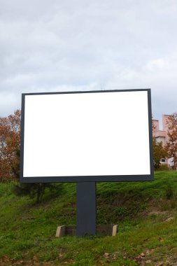Yeşil bir kentsel alanda duran, bulutlu bir günde çekilmiş büyük boş bir ilan panosu. Boş beyaz reklam paneli çimenler, ağaçlar ve sonbahar yapraklarıyla çevrili koyu metal bir yapının üzerine monte edilmiştir. İdeal model