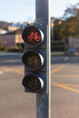 Şehir kavşağındaki bir direğe monte edilmiş kırmızı ışığı gösteren bir bisiklet trafik ışığının yakın çekimi. Görüntü karayolu güvenliği, bisiklet altyapısı, sürdürülebilir ulaşım ve şehir trafik kontrolünü temsil ediyor.