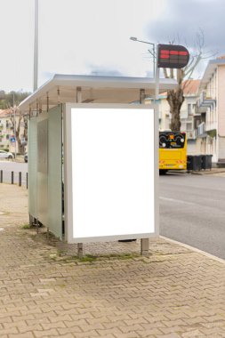 Sakin bir şehir caddesi boyunca bir otobüs durağı barınağında boş bir reklam panosu modeli. Boş reklam paneli, arka planda konut binaları ve trafik ışıkları bulunan markalaşma ve pazarlama kampanyaları için temiz bir kopya alanı sunuyor..