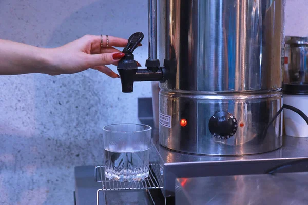 Une main de femme presse le robinet pour verser de l'eau chaude de la chaudière dans un verre transparent sur la table — Photo