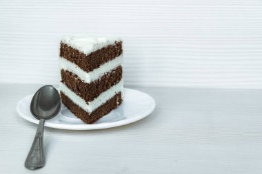 Sol tarafta tatlı kaşığı ve tereyağı kremalı bir parça çikolatalı kek var.