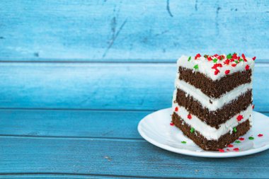 Sağında tereyağı kremi olan bir dilim çikolatalı kek. Mavi arka plan. Kırmızı-yeşil toz alma. Yakın plan..