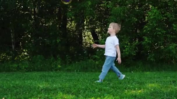 Joyeux petit garçon aux cheveux blonds catc grosses bulles de savon dans le parc d'été. Rire et courir en gros plan enfant en t-shirt blanc. HD 1920 1080. Ralentissez 50 ips. extérieur. — Video