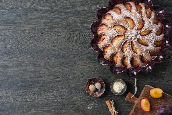 Pflaumenkuchen und Zutaten auf dunklem Holztisch — Stockfoto