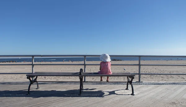 Little girl sitting on the bench looking out at the ocean — Stock fotografie