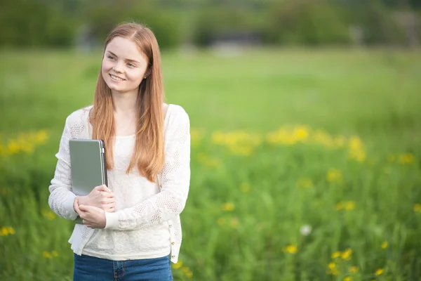 Gelukkig meisje Europees uiterlijk staande op het gras met een laptop op groene natuur achtergrond — Stockfoto