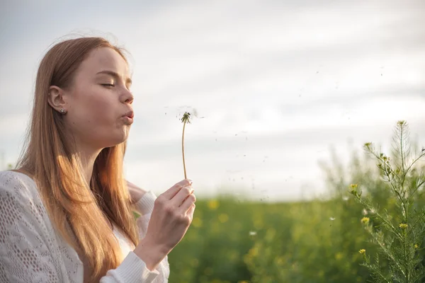 Junge Frühlingsmode-Frau bläst Löwenzahn im Frühlingsgarten. Frühling. Trendiges Mädchen bei Sonnenuntergang im Frühling Landschaft Hintergrund. Allergisch gegen Blütenpollen. Frühlingsallergie. — Stockfoto