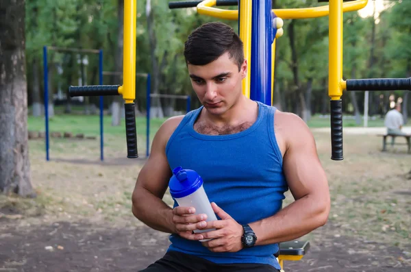 Imagen del guapo deportista de camisa azul con batido de proteínas en el parque con gimnasio . — Foto de Stock