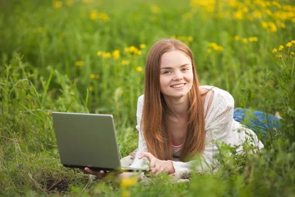 Wanita muda tersenyum berbaring di rumput dengan notebook di taman mencari kamera . — Stok Foto