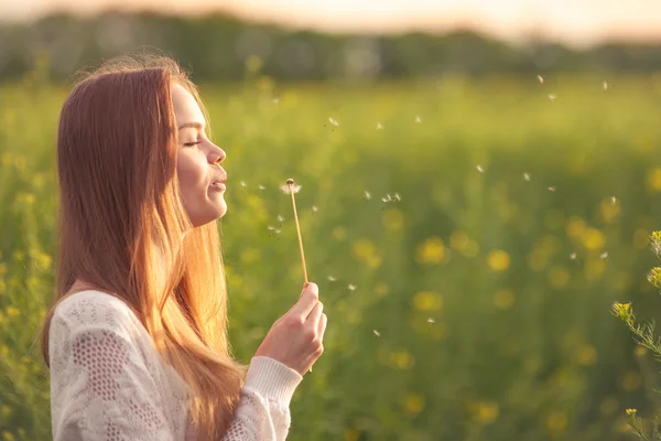 Jeune femme de mode de printemps soufflant le pissenlit dans le jardin de printemps. Au printemps. Fille à la mode au coucher du soleil dans le paysage de printemps fond. Allergique au pollen des fleurs. Allergie de printemps. — Photo