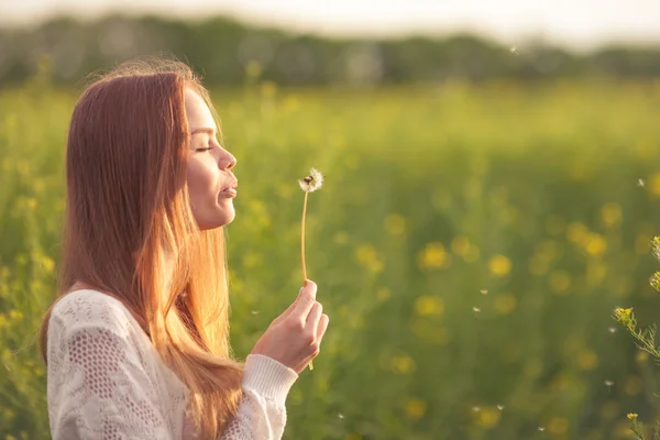 Junge Frühlingsmode-Frau bläst Löwenzahn im Frühlingsgarten. Frühling. Trendiges Mädchen bei Sonnenuntergang im Frühling Landschaft Hintergrund. Allergisch gegen Blütenpollen. Frühlingsallergie. — Stockfoto