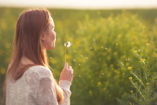 Młody wiosna moda kobieta dmuchanie mniszka lekarskiego w spring garden. Sezon wiosenny. Modny dziewczyna o zachodzie słońca w tle pejzaż wiosna. — Zdjęcie stockowe