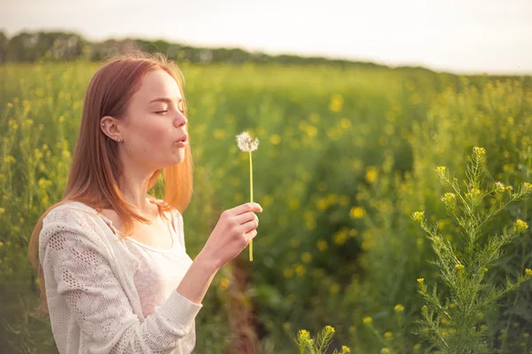 Joven mujer de la moda de primavera soplando diente de león en el jardín de primavera. Primavera. Chica de moda al atardecer en el fondo del paisaje de primavera —  Fotos de Stock